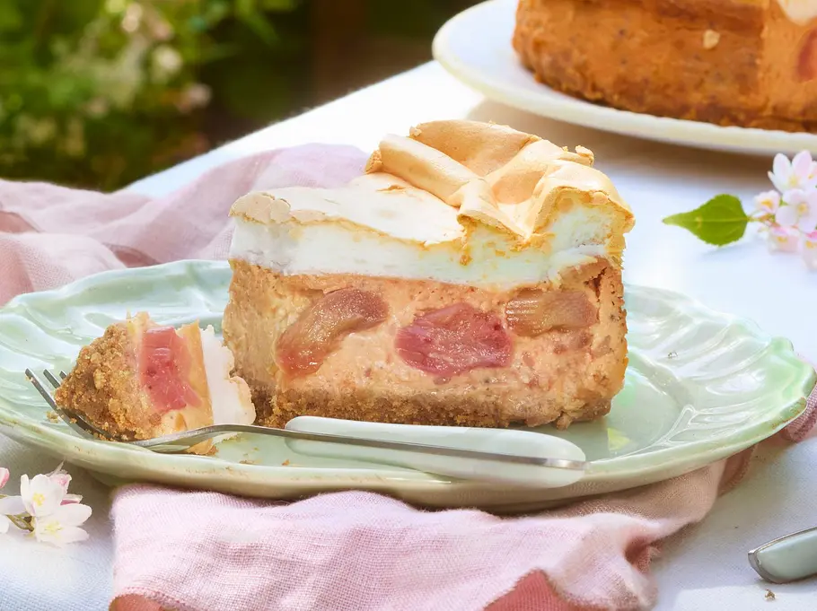 Erdbeer-Rhabarber-Baiserkuchen mit goldbraun gebackenem Boden, saftiger Fruchtschicht aus Erdbeeren und Rhabarber und einer luftig gebräunten Baiserhaube. Ein Stück liegt auf einem zarten Pastellteller mit Gabel, der restliche Kuchen steht auf einer Tortenplatte daneben.