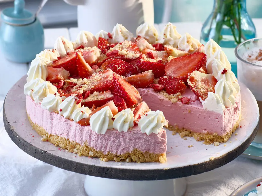 Erdbeer-Skyr-Cheesecake mit rosa Creme, frischen Erdbeerstückchen und Sahnetupfen auf Keksboden, serviert auf einer weißen Kuchenplatte.