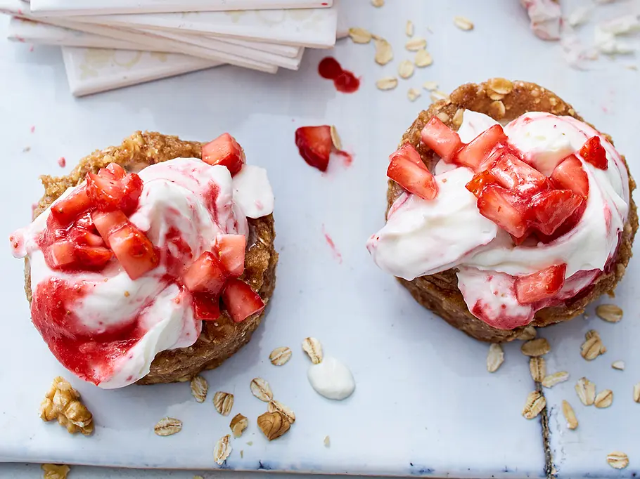 Erdbeer-Skyr-Törtchen ohne Zucker Rezept Zwei kleine Törtchen mit Skyr-Creme und frischen Erdbeerstücken, auf einer hellen Platte mit Haferflocken dekoriert.