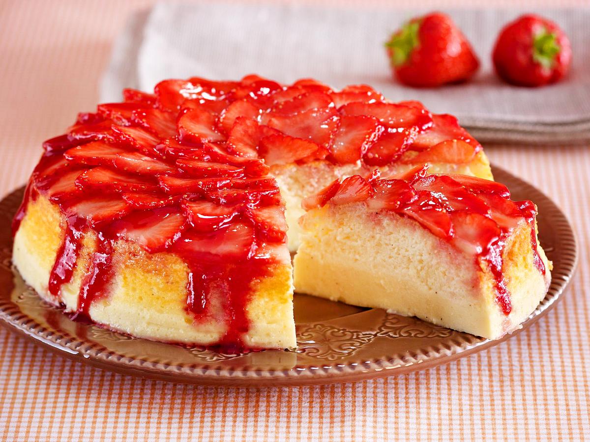 Der in einer Springform gebackene Erdbeer-Soufflé-Käsekuchen ist  zu einem Viertel angeschnitten. Der Anschnitt ist zu erkennen. Unten wirkt der Teig kompakt wie Käsekuchen, darauf ist eine luftige Kuchenschicht zu erkennen. Getoppt wird der Kuchen von frischen Erdbeerscheiben und etwas Konfitüre, die herunterläuft.