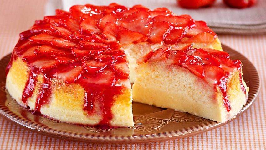 Der in einer Springform gebackene Erdbeer-Soufflé-Käsekuchen ist  zu einem Viertel angeschnitten. Der Anschnitt ist zu erkennen. Unten wirkt der Teig kompakt wie Käsekuchen, darauf ist eine luftige Kuchenschicht zu erkennen. Getoppt wird der Kuchen von frischen Erdbeerscheiben und etwas Konfitüre, die herunterläuft. - Foto: House of Food / Bauer Food Experts KG