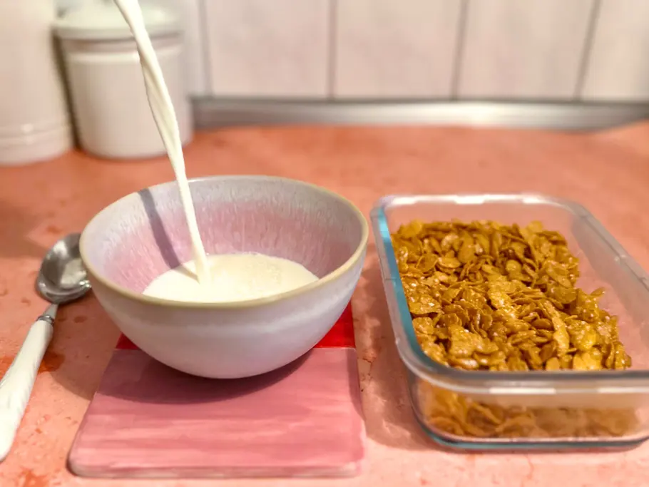 Milch wird in eine Müslischale gegossen, daneben stehen eiskalte Cornflakes in einer Glasschale auf einem rosa Untergrund