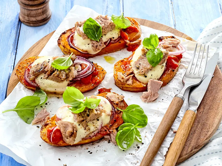 Süßkartoffel-Pizzette al tonno Rezept Süßkartoffel-Pizzette mit Tomatensauce, geschmolzenem Käse, Thunfisch und roten Zwiebeln, mit frischem Basilikum belegt und auf einem Holzbrett serviert.
