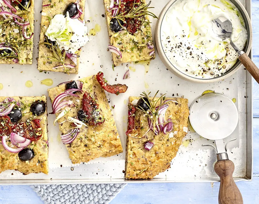 In Stücke geschnittene Low-Carb-Focaccia mit Oliven, roten Zwiebeln, getrockneten Tomaten und Rosmarin, auf einem Blech angerichtet, dazu ein Schälchen Dip.