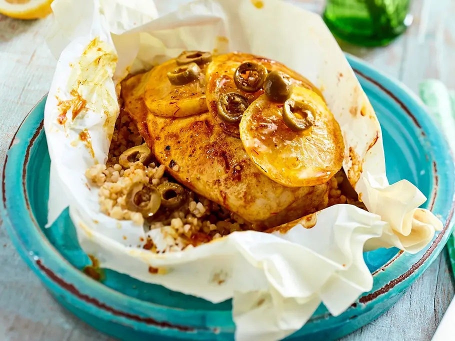 Saftiges Hähnchenfilet mit Zitronenscheiben und grünen Oliven, serviert auf lockerem Couscous in Backpapier, angerichtet auf einem türkisfarbenen Teller.