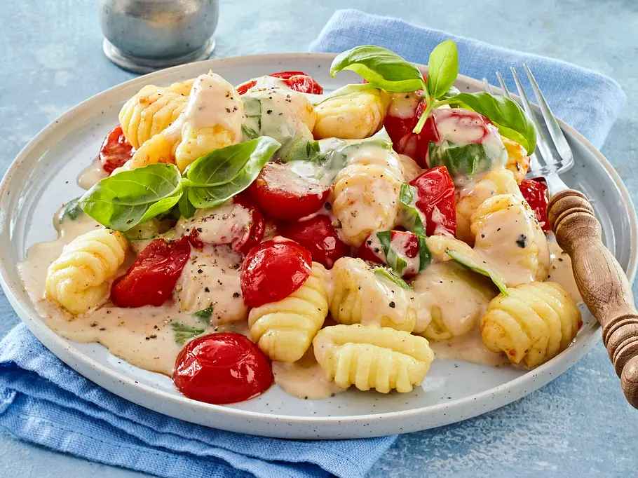 Cremige Gnocchi mit Cocktailtomaten, frischem Basilikum und heller Soße auf einem weißen Teller, serviert auf einem blauen Stofftuch.