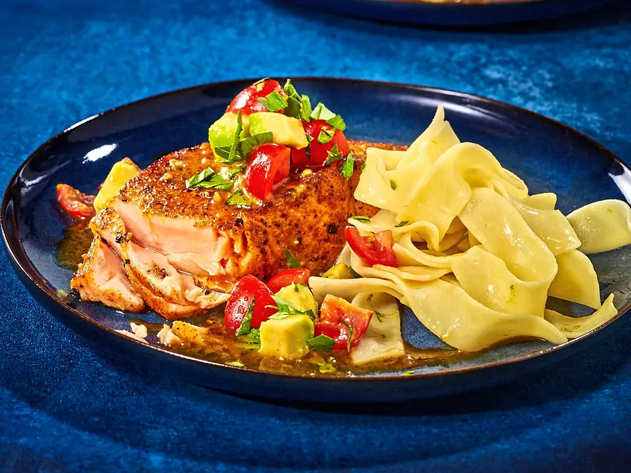 Cajun-Lachsfilets mit Bandnudeln Zartes Cajun-Lachsfilet auf einem dunklen Teller, dazu breite Bandnudeln und Würfel von Tomaten und Avocado als frische Garnitur.