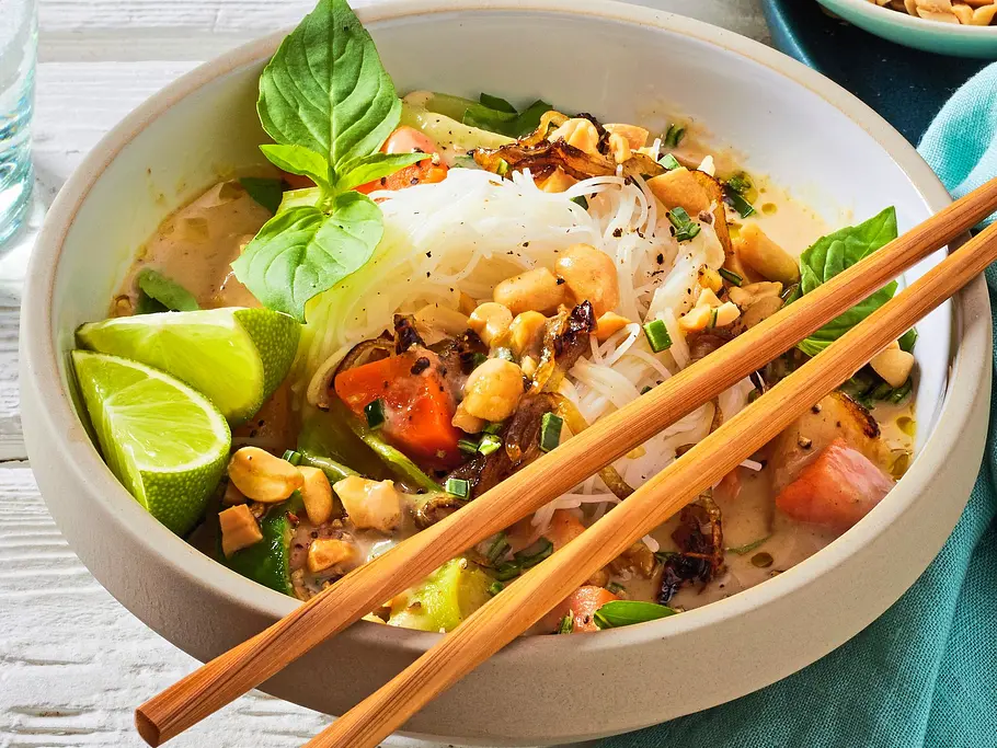 Bunte Bowl mit Reisnudeln, knackigem Rüben-Mix, gehackten Erdnüssen, Limetten und frischem Basilikum in einer hellen Schale, dazu Essstäbchen auf einem weißen Holztisch.