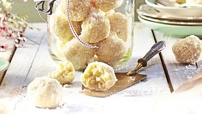 Kokos-Trüffel in runder Form, mit Kokosraspeln umhüllt, dekorativ in einem Glas serviert und einige einzeln und angeschnitten auf einem hellen Holztisch platziert. - Foto: LECKER @ Bauer Media Group
