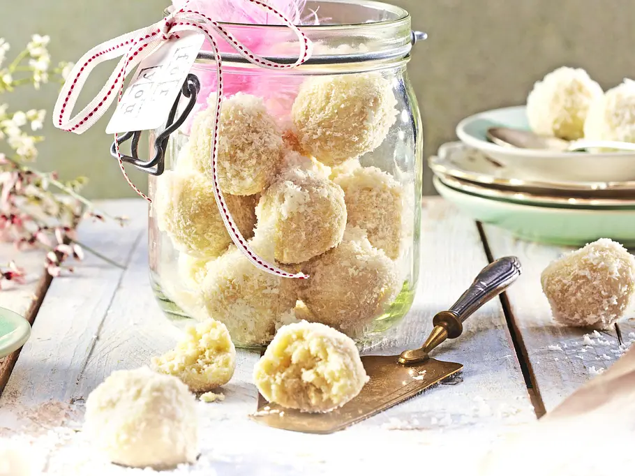 Kokos-Trüffel in runder Form, mit Kokosraspeln umhüllt, dekorativ in einem Glas serviert und einige einzeln und angeschnitten auf einem hellen Holztisch platziert.