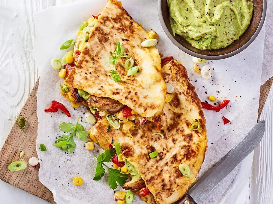 Knusprig gebratene Quesadilla mit Hähnchen, Paprika, Mais und Frühlingszwiebeln auf einem Holzbrett, daneben ein Schälchen Guacamole.
