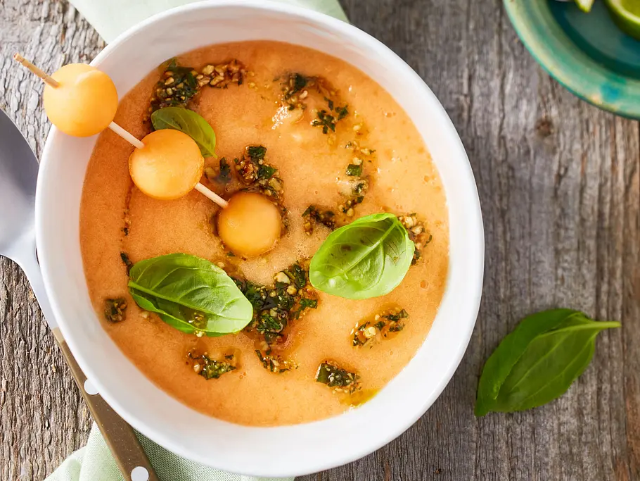 Kalte Melonensuppe mit Basilikumpesto, garniert mit Melonenkugeln am Spieß und frischem Basilikum, serviert in einer weißen Schale auf rustikalem Holztisch.