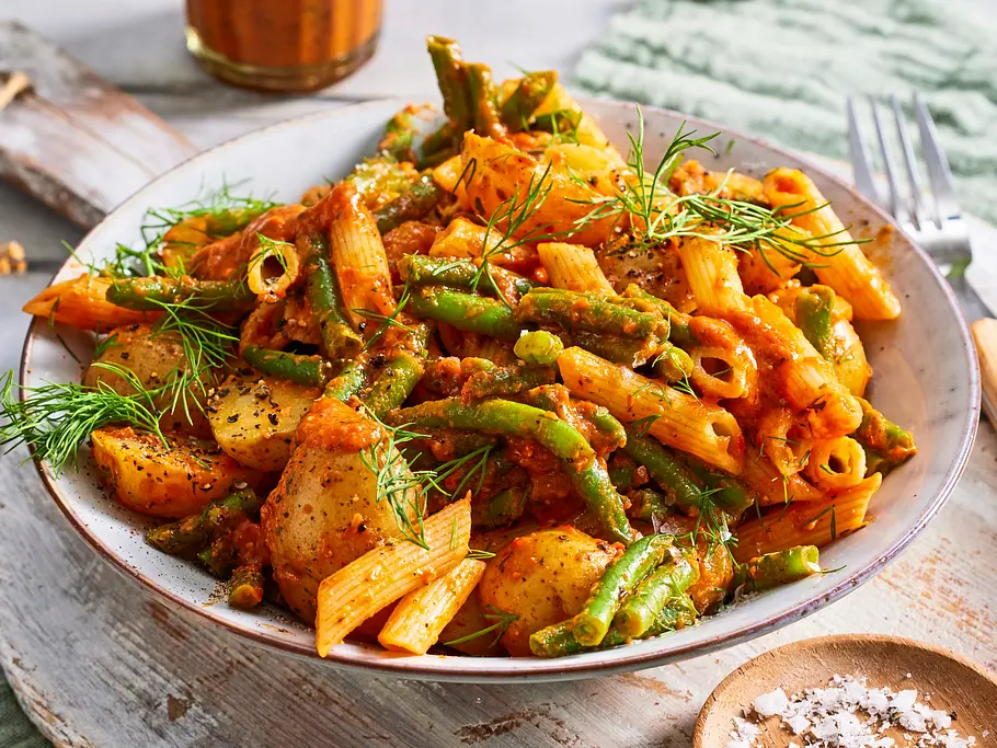 Pasta mit grünen Bohnen und Kartoffelstücken in rotem Pesto, serviert auf einem weißen Teller mit frischem Dill, auf rustikalem Holzbrett und grünem Stoff.