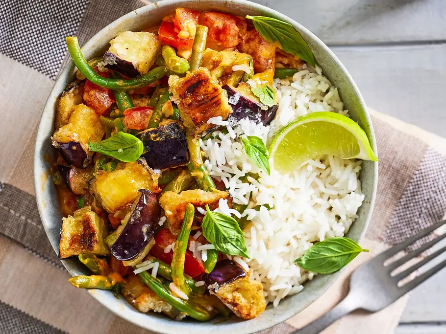 Sommerliches Auberginen-Bohnen-Curry Schüssel mit buntem Sommergemüse-Curry und Basmatireis, garniert mit frischen Basilikumblättern und einer Limettenspalte, auf einem hellen Keramikteller und beiger Stoffserviette angerichtet.