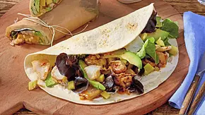 Soft-Wraps gefüllt mit knusprigem Fisch, Salat und Avocado, auf einem runden Holzbrett serviert. Daneben Schälchen mit Dip und Mango sowie ein Glas Wasser. - Foto: LECKER @ Bauer Media Group