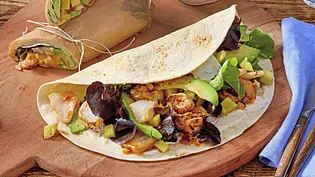 Soft-Wraps gefüllt mit knusprigem Fisch, Salat und Avocado, auf einem runden Holzbrett serviert. Daneben Schälchen mit Dip und Mango sowie ein Glas Wasser. - Foto: LECKER @ Bauer Media Group