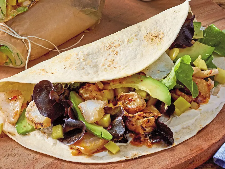 Soft-Wraps gefüllt mit knusprigem Fisch, Salat und Avocado, auf einem runden Holzbrett serviert. Daneben Schälchen mit Dip und Mango sowie ein Glas Wasser.