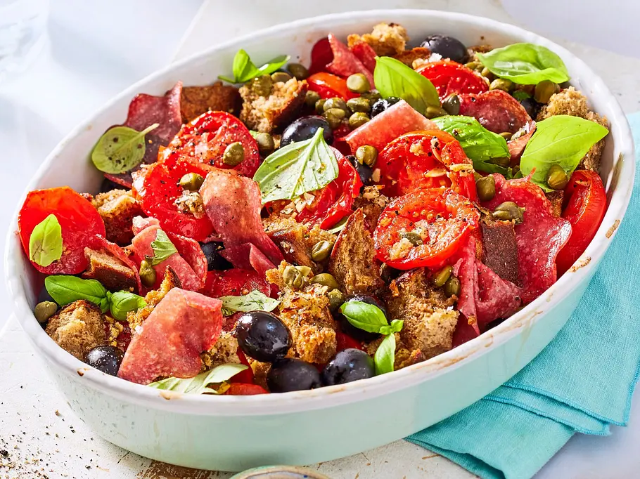 Warmer Tomaten-Brot-Salat mit Oliven Rezept Warmer Tomaten-Brot-Salat mit schwarzen Oliven, Kapern, knusprigen Brotwürfeln und frischem Basilikum, serviert in einer hellen Schale auf einem weißen Brett.