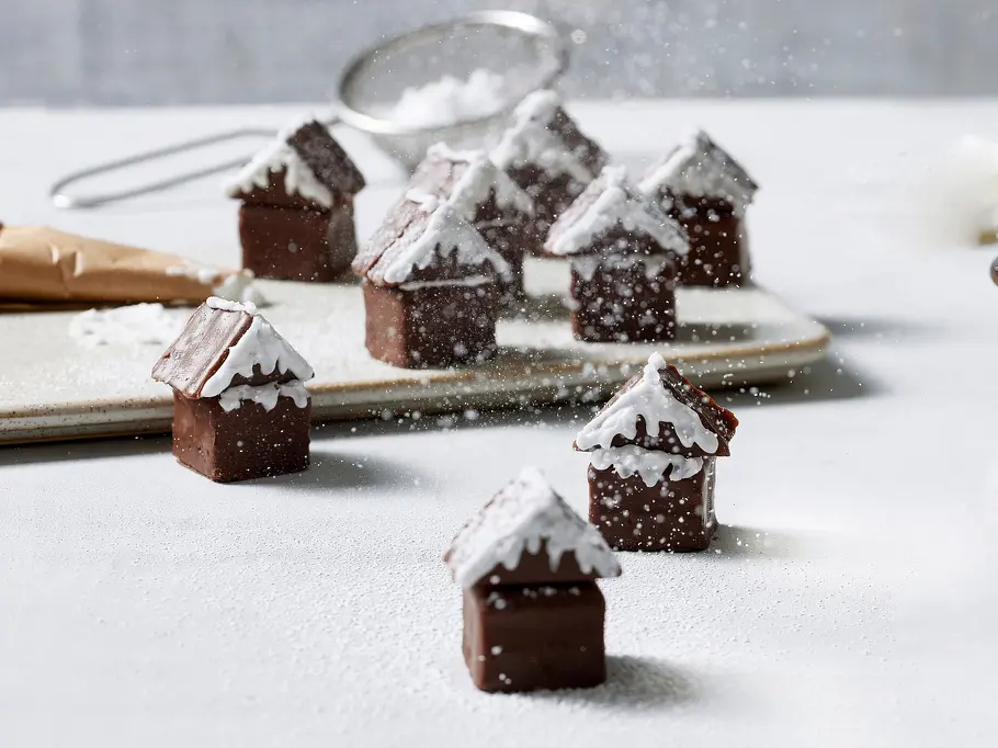 Kleine Winter-Häuschen Rezept Kleine Winter-Häuschen aus Schokoladenwürfeln, zusammengebaut und mit weißem Zuckerguss als Schnee dekoriert, im Hintergrund weitere Schoko-Konfekte und ein Glas Schokoladencreme.