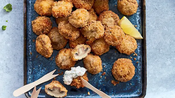 Knusprige, goldbraun panierte Champignons auf einem blauen Tablett, dazu ein Schälchen cremiger Dip mit Kräutern und einer Zitronenspalte, garniert mit frischer Minze. Einige der Champignons sind bereits angeschnitten und mit Dip serviert. - Foto: LECKER @ Bauer Media Group Knusprige, goldbraun panierte Champignons auf einem blauen Tablett, dazu ein Schälchen cremiger Dip mit Kräutern und einer Zitronenspalte, garniert mit frischer Minze. Einige der Champignons sind bereits angeschnitten und mit Dip serviert. - Foto: LECKER @ Bauer Media Group