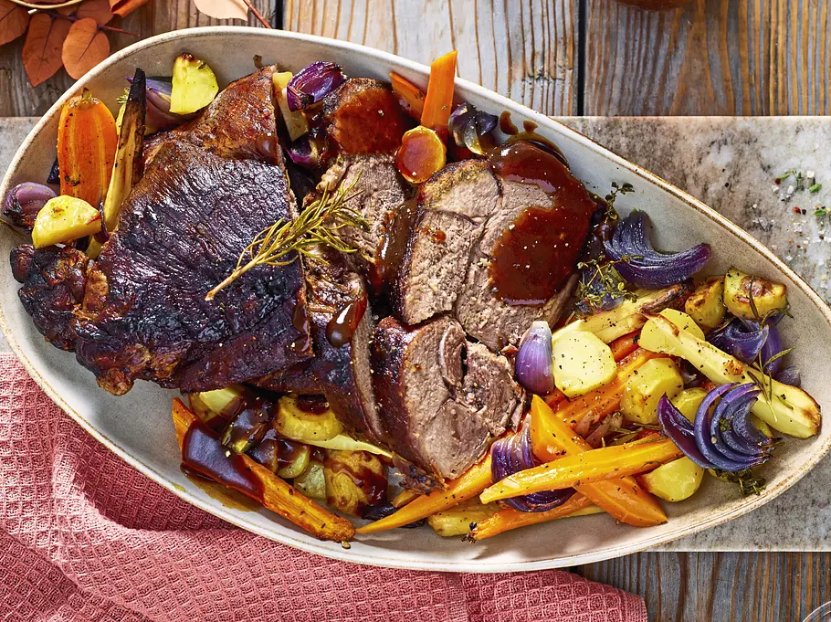 Lammkeule in Rotwein-Cassis-Soße Rezept Scheiben einer geschmorten Lammkeule mit dunkler Rotwein-Cassis-Soße auf ovaler Keramikplatte, dazu bunte Ofenkartoffeln, Karotten und rote Zwiebeln.