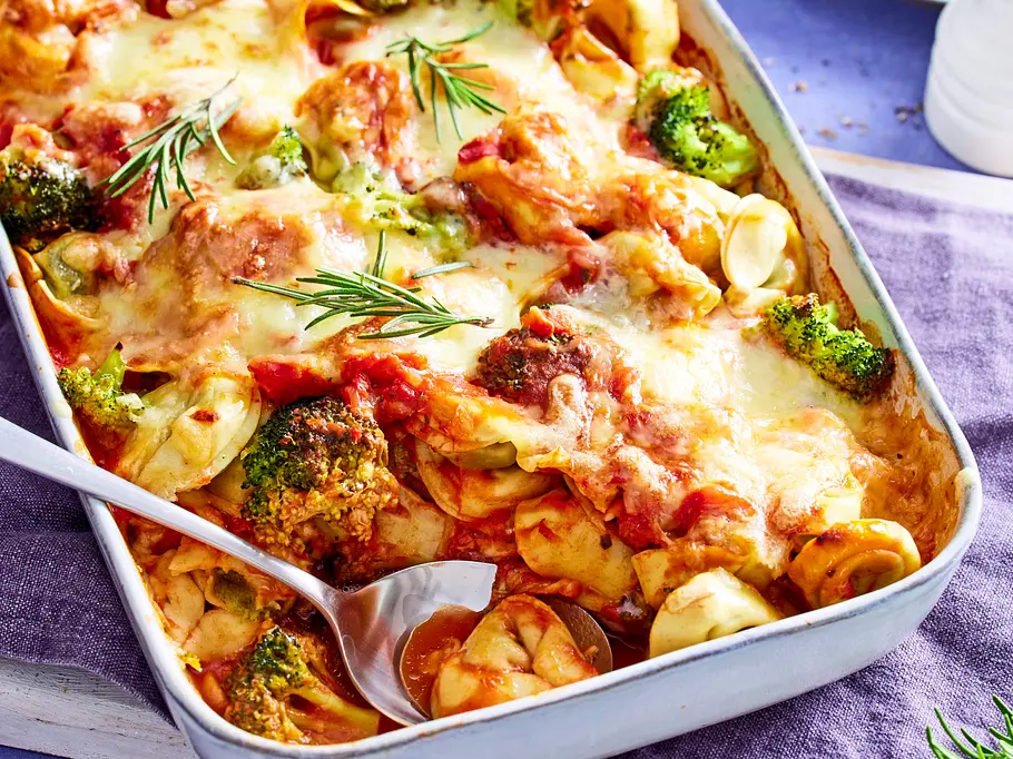 Tortelloni-Brokkoli-Auflauf mit viel geschmolzenem Käse und Tomatensauce in einer Auflaufform, garniert mit Rosmarin, auf einer violetten Stoffserviette serviert.