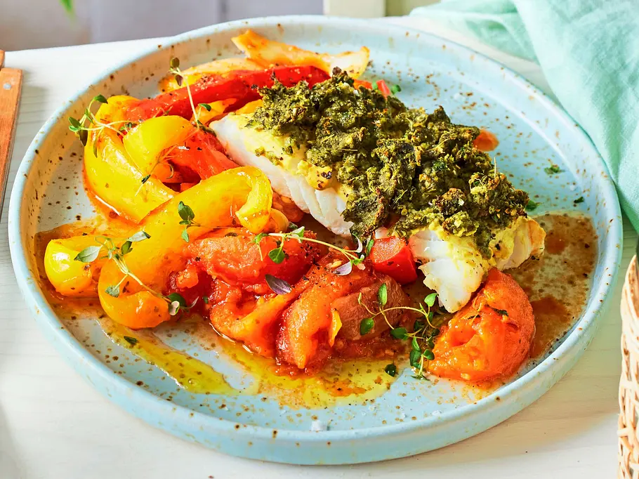 Kabeljaufilet mit Kräuterkruste auf einem Bett aus bunt geschmortem Paprikagemüse, serviert auf einem hellblauen, gesprenkelten Keramikteller.