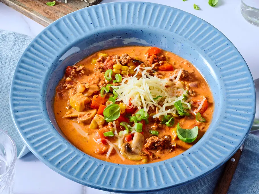 Cremige Bolognese-Suppe Cremige, orangefarbene Bolognese-Suppe mit Hackfleisch, Gemüse und geriebenem Käse, angerichtet in einem tiefen, blauen Teller auf hellblauer Stoffserviette.