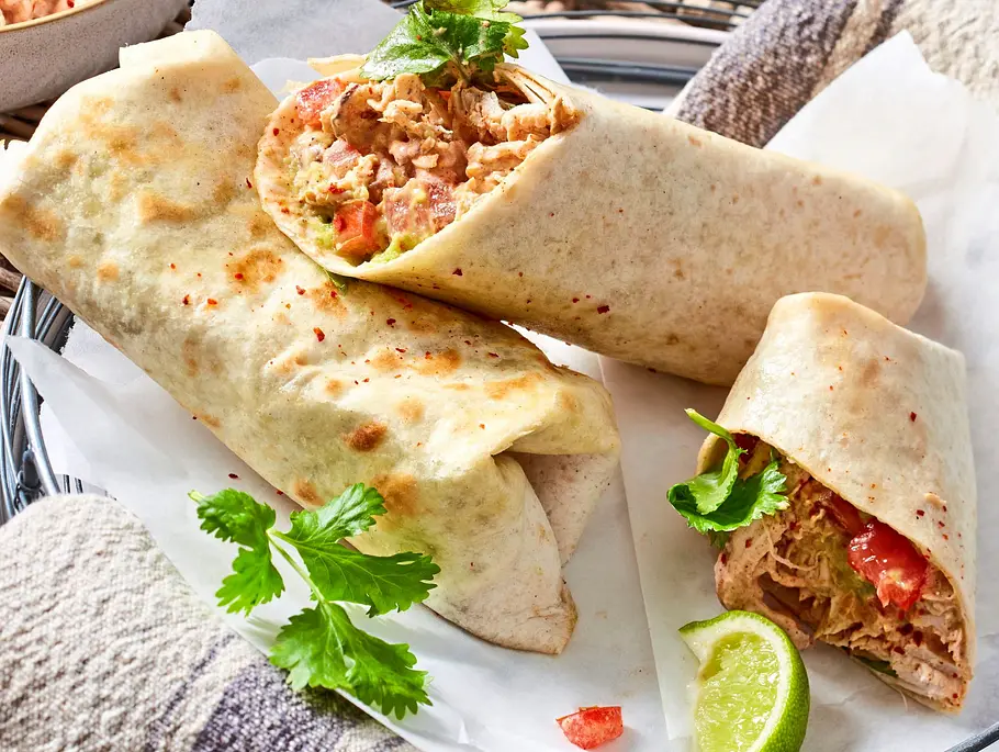 Aufgeschnittene Pulled-Chicken-Burritos auf einem weißen Teller, gefüllt mit zartem Hähnchen, Gemüse und frischen Kräutern, dekoriert mit Limettenspalten und einem Tuch als Unterlage.