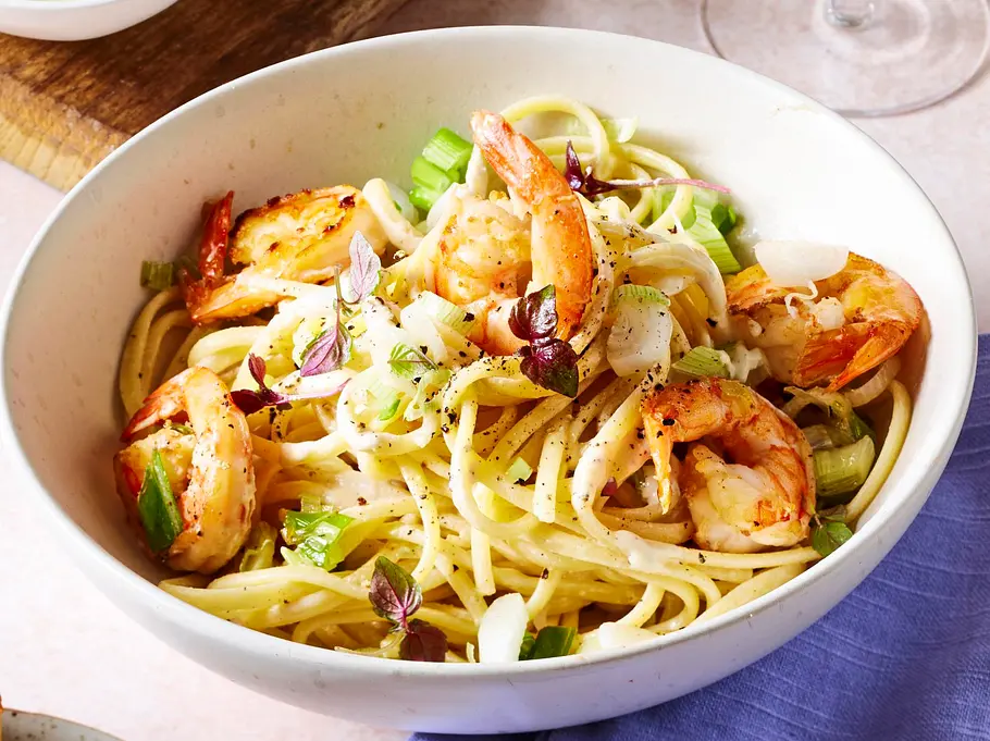 Spaghetti mit gebratenen Garnelen und Frühlingszwiebeln in einer weißen Schale, dazu zwei Scheiben knuspriges Knoblauchbrot auf einem kleinen Teller, serviert auf einer hellen, glatten Unterlage mit einem lilafarbenen Tuch darunter.