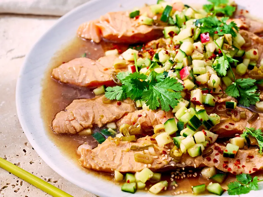 Heiß marinierter Lachs mit Gurken-Apfel-Mix Saftige Lachsscheiben in einer klaren Marinade, garniert mit würzigem Gurken-Apfel-Mix und frischem Koriander, serviert auf einem weißen Teller.