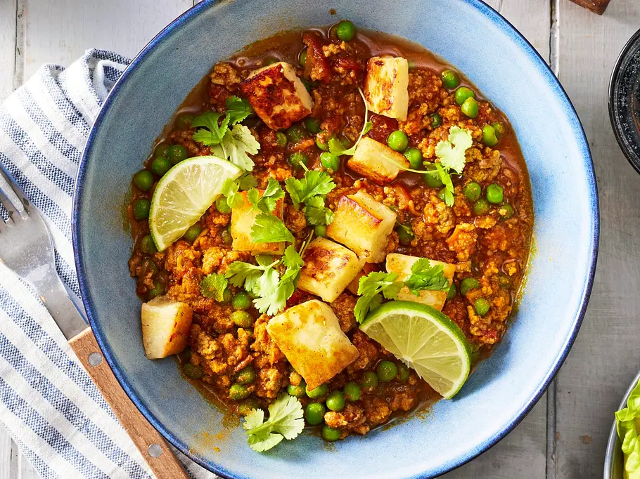 Halloumi-Hack-Pfanne Rezepte Halloumi-Hack-Pfanne mit würzigem Hackfleisch, grünen Erbsen und goldbraun gebratenen Halloumi-Würfeln, verfeinert mit Koriander und Limettenspalten, serviert in einer Pfanne und in einer blauen Schale mit Holzlöffel.