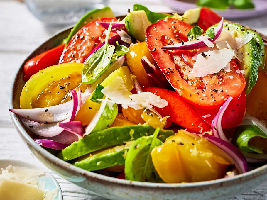 Avocado-Summer-Bowl Bunte Bowl mit Avocado, Tomaten in verschiedenen Farben, roten Zwiebeln, frischem Basilikum und Parmesanspänen, serviert in einer hellen Keramikschale.