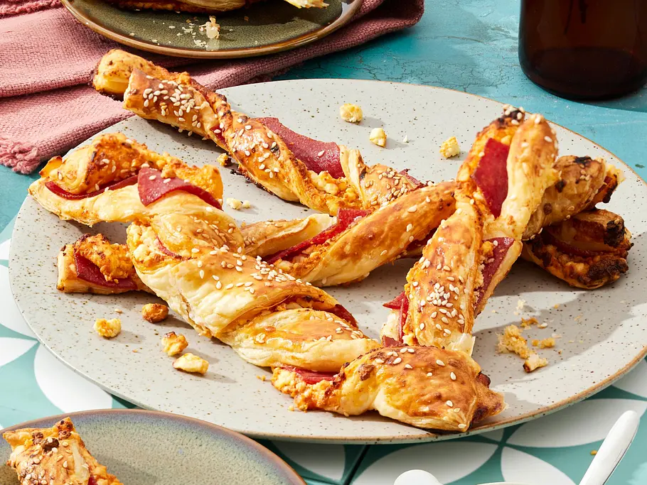 Salami-Käse-Stangen aus dem Airfryer Mehrere goldbraune Blätterteig-Stangen mit Salami und geschmolzenem Käse, bestreut mit Sesam, auf rustikalen Tellern serviert. Daneben eine kleine Schale mit Dip.