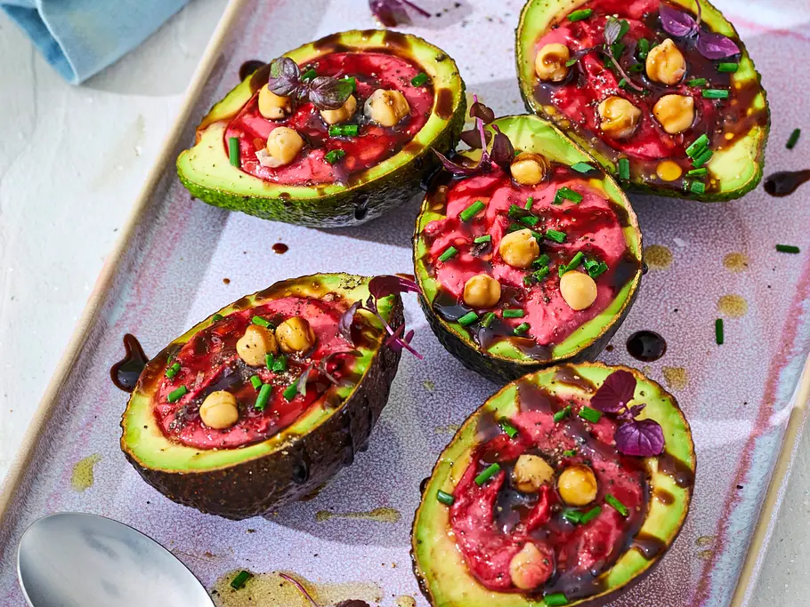 Halbierte Avocados, gefüllt mit leuchtend pinkem Rote-Bete-Hummus, garniert mit Kichererbsen, frischen Kräutern und etwas Balsamico, angerichtet auf einer hellen, rechteckigen Servierplatte. Daneben liegen zwei Löffel auf der Platte.