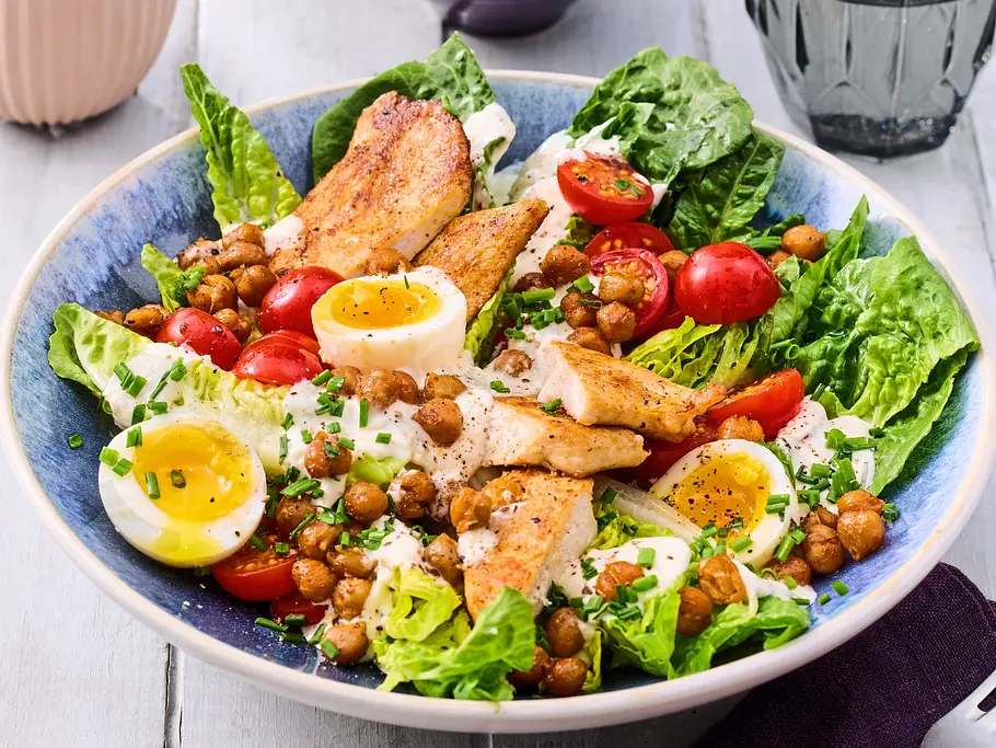 Bunte Bowl mit knackigem Römersalat, gebratenen Hähnchenstreifen, Kichererbsen, halbierten Kirschtomaten, weichen gekochten Eiern und cremigem Dressing, angerichtet auf einem blauen Keramikteller auf hellem Holztisch.
