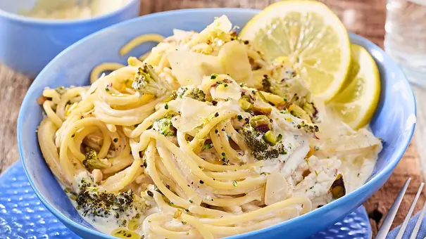 Spaghetti in einer cremigen Zitronensoße mit Brokkoliröschen, gerösteten Pistazien und gehobeltem Parmesan, garniert mit frischen Zitronenscheiben, serviert in einer hellblauen Schale auf einem strukturierten blauen Teller. - Foto: LECKER @ Bauer Media Group