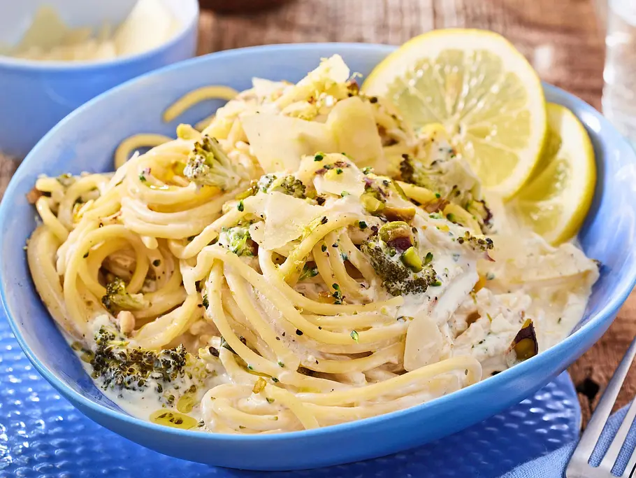 Spaghetti in einer cremigen Zitronensoße mit Brokkoliröschen, gerösteten Pistazien und gehobeltem Parmesan, garniert mit frischen Zitronenscheiben, serviert in einer hellblauen Schale auf einem strukturierten blauen Teller.