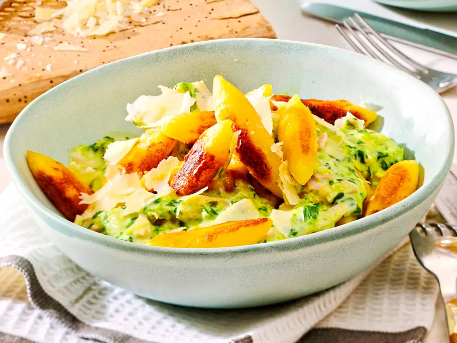 Knusprig gebratene Schupfnudeln in einer cremigen Sahnesoße mit Spinat und Schinken, garniert mit gehobeltem Parmesan, serviert in einer mintgrünen Schüssel.