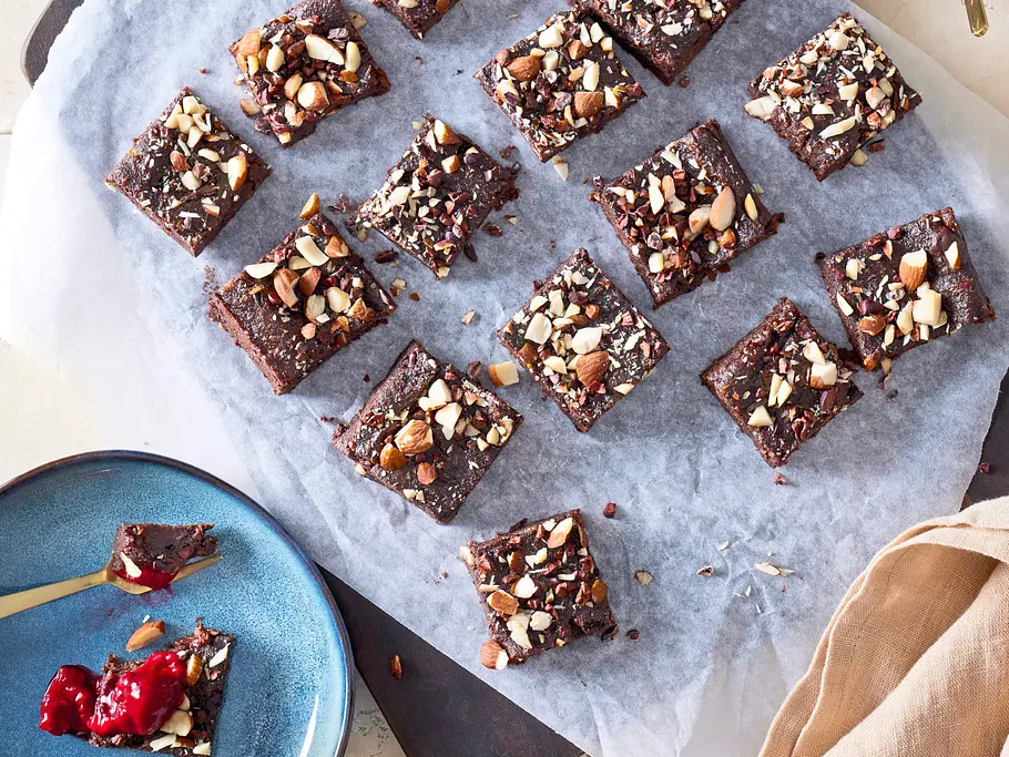 Kleine, eckige Brownie-Stücke mit gehackten Nüssen bestreut, auf einem weißen Backpapier und dunklem Holzbrett angerichtet. Daneben eine blaue Schale mit rotem Fruchtkompott und ein blauer Teller mit einem Brownie-Stück und Fruchtaufstrich.