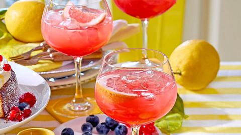 Glas Campari Amalfi mit leuchtend rotem Drink auf Eis, garniert mit einer Grapefruitscheibe, auf einem gelb-weiß gestreiften Tischdecko mit Zitronen im Hintergrund. - Foto: Image Professionals