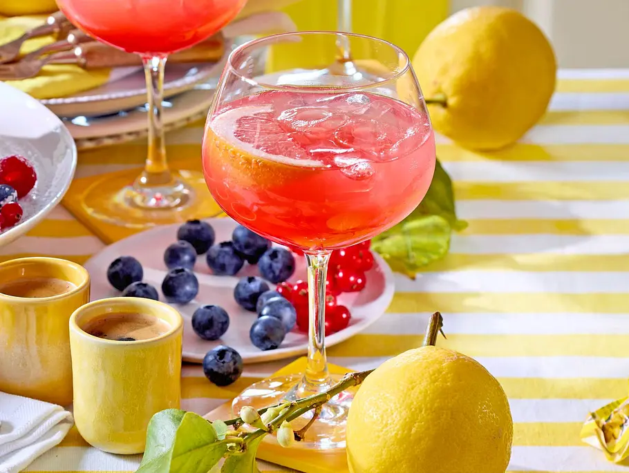Campari Amalfi Rezept Glas Campari Amalfi mit leuchtend rotem Drink auf Eis, garniert mit einer Grapefruitscheibe, auf einem gelb-weiß gestreiften Tischdecko mit Zitronen im Hintergrund.