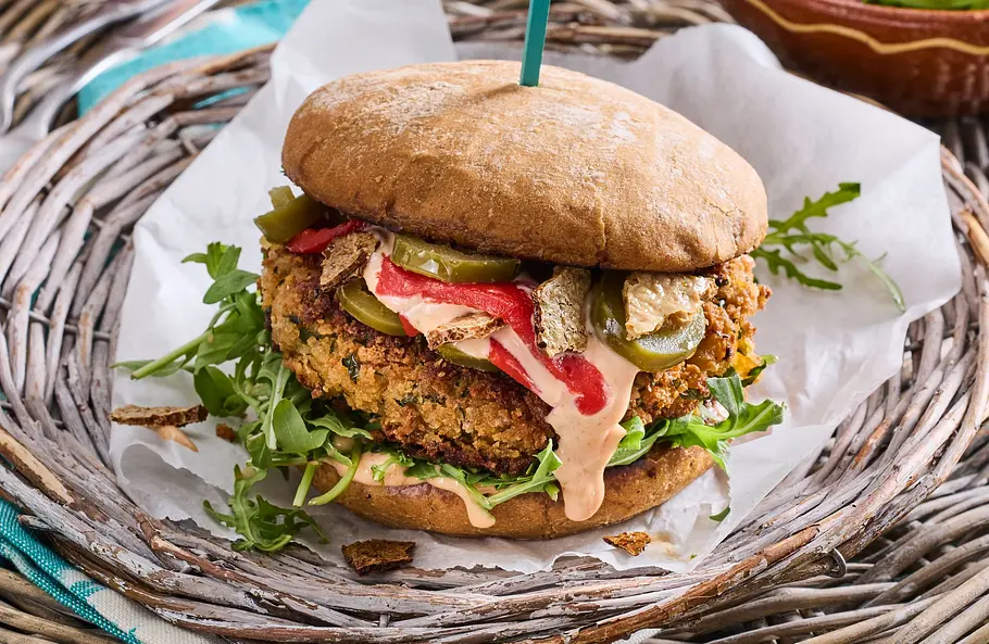 Falafel-Burger mit Tofu-Bacon Falafel-Burger im Brötchen mit Rucola, Sauce und Tofu-Bacon, angerichtet auf weißem Backpapier auf einem runden Flechttablett.