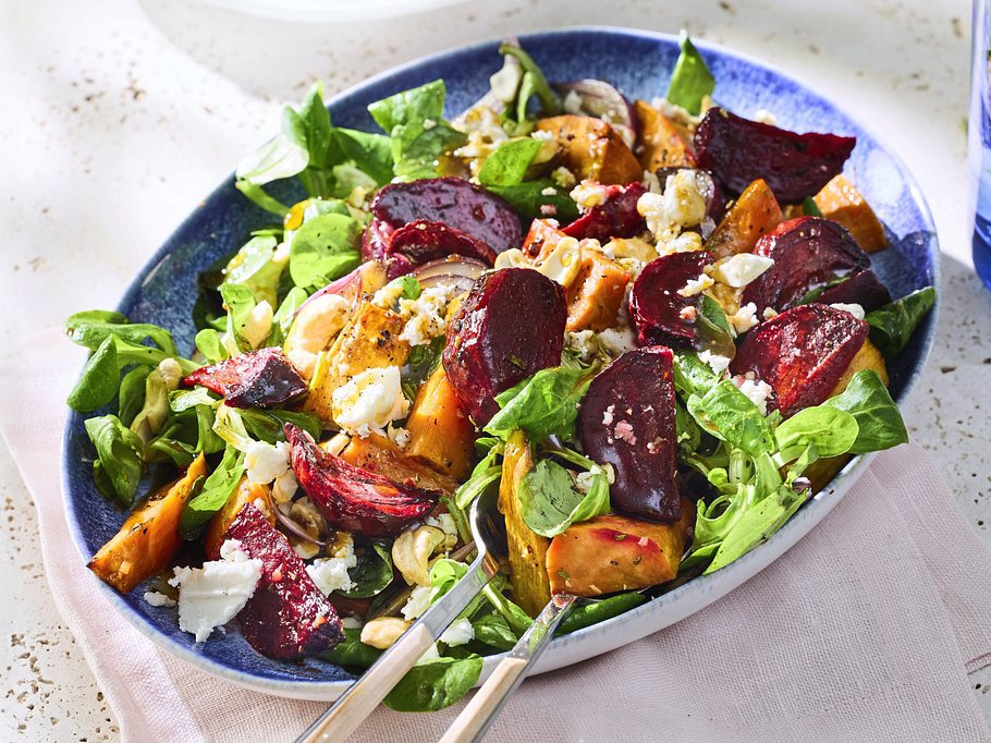 Bunter Salat aus lauwarmer, gerösteter Rote Bete und Süßkartoffeln auf frischem Blattgrün, garniert mit zerbröseltem Feta und Cashewkernen, serviert auf einem dunkelblauen Teller.