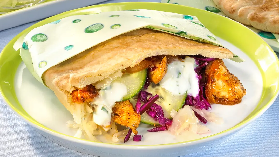 Gefüllte Pita-Tasche mit Hähnchenstücken, Rotkohl, Gurken, Krautsalat und Joghurtsauce auf grün-weißem Teller, daneben weitere Pitabrote und kleine Schalen mit Krautsalat und Sauce. - Foto: LECKER @ Bauer Media Group