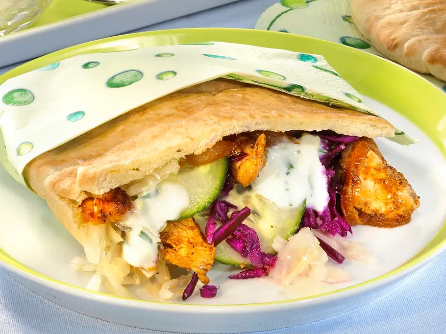 Gefüllte Pita-Tasche mit Hähnchenstücken, Rotkohl, Gurken, Krautsalat und Joghurtsauce auf grün-weißem Teller, daneben weitere Pitabrote und kleine Schalen mit Krautsalat und Sauce.