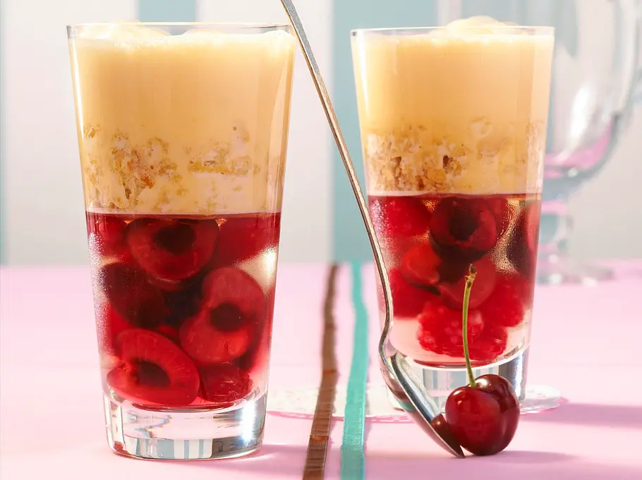 boozy Kirsch-Trifle Zwei Gläser mit Schichtdessert aus Kirschen, Kirschgelee, zerbröseltem Biskuit und gelber Creme auf einem rosa Tisch, ein langer Löffel und eine Kirsche als Dekoration.