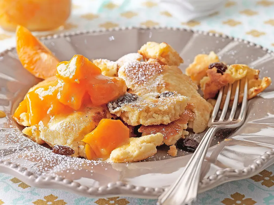 Locker zerzupfter Kaiserschmarrn mit Rosinen, bestäubt mit Puderzucker, auf einem verzierten grauen Teller, dazu leuchtend gelbes Kompott und Gabel.