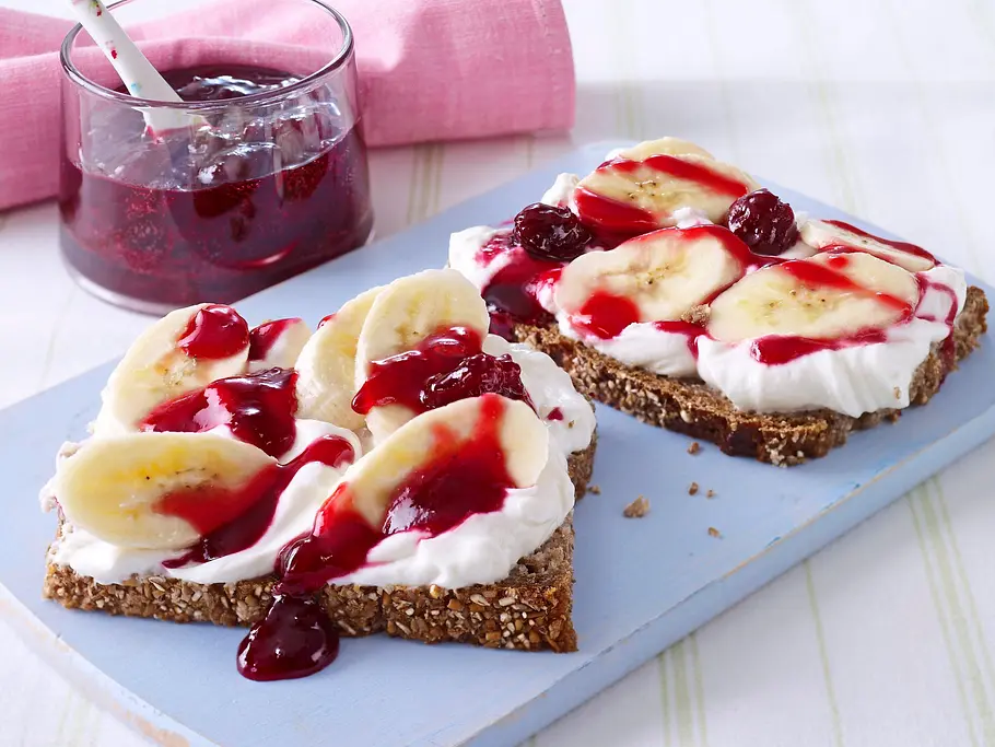 Zwei Scheiben Körnerbrot mit Quark, frischen Bananenscheiben und Kirschmarmelade auf einem hellblauen Holzbrett serviert.