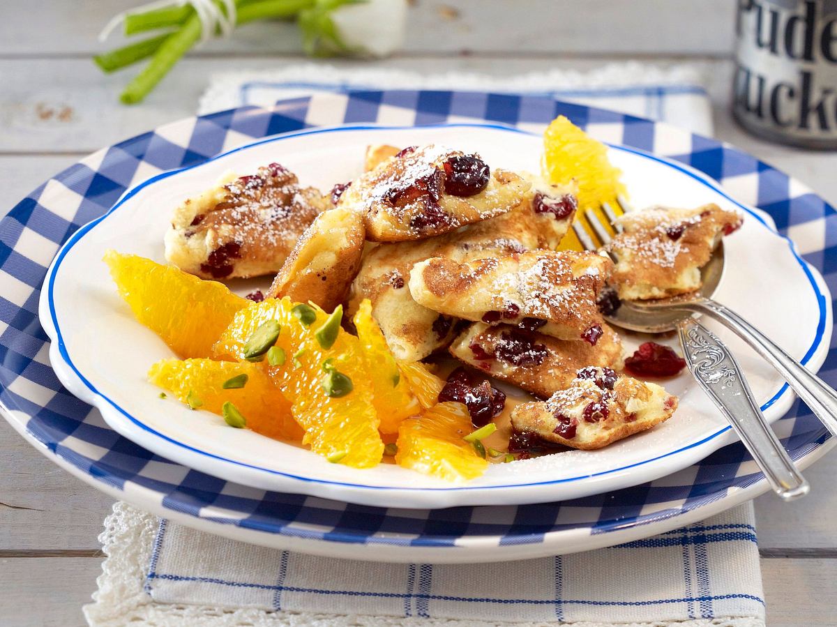 Cranberry-Schmarren, bestäubt mit Puderzucker, serviert mit heißen Orangenfilets und Pistazien auf einem karierten Teller.